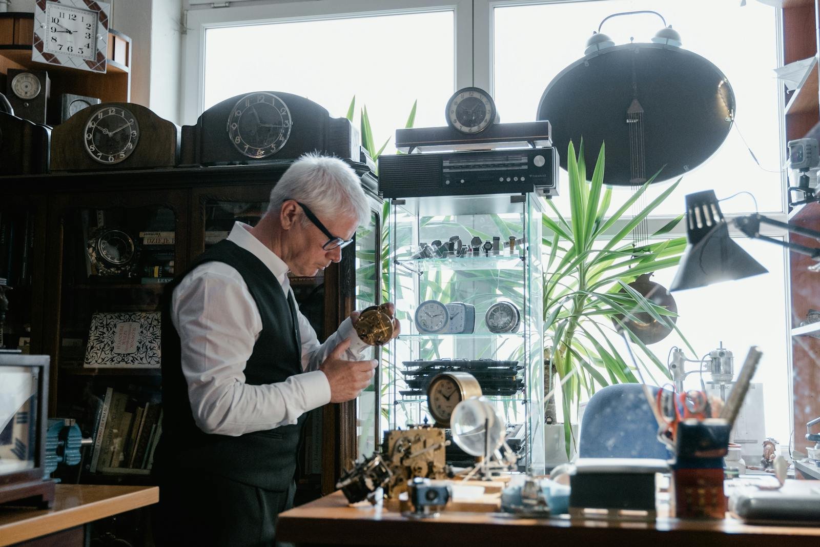 Elderly man repairing clocks in a workshop surrounded by timepieces and tools.